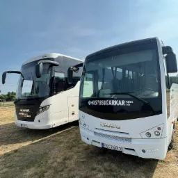 Dwa białe autokary, Scania i Isuzu, firmy przewozowej z Nowego Tomyśla, stojące na trawiastym placu w słoneczny dzień. Na szybie Isuzu widnieje numer telefonu firmy i napis 'Przewóz osób'.