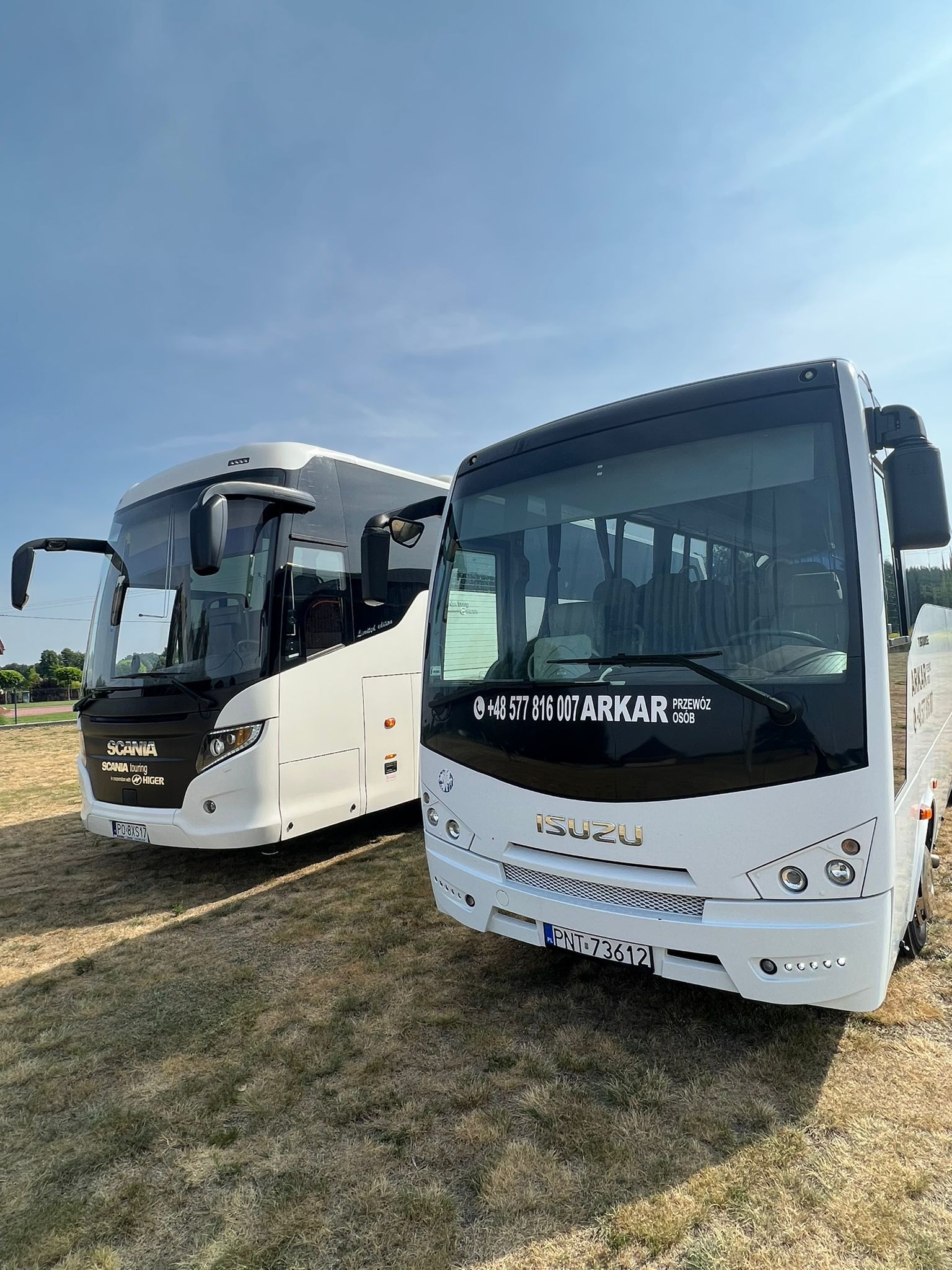 Dwa białe autokary, Scania i Isuzu, firmy przewozowej z Nowego Tomyśla, stojące na trawiastym placu w słoneczny dzień. Na szybie Isuzu widnieje numer telefonu firmy i napis 'Przewóz osób'.