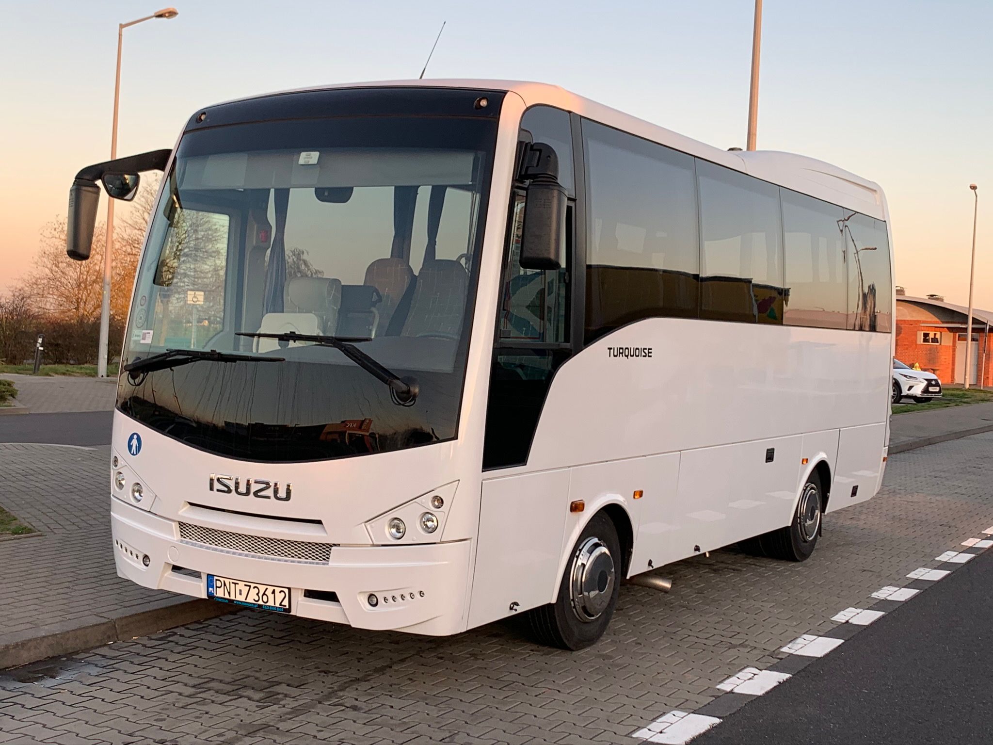 Biały autobus marki Isuzu Turquoise zaparkowany na kostce brukowej, z widocznym znakiem osoby niepełnosprawnej na przedniej szybie i numerem rejestracyjnym PNT 73612, sfotografowany w słoneczny dzień.