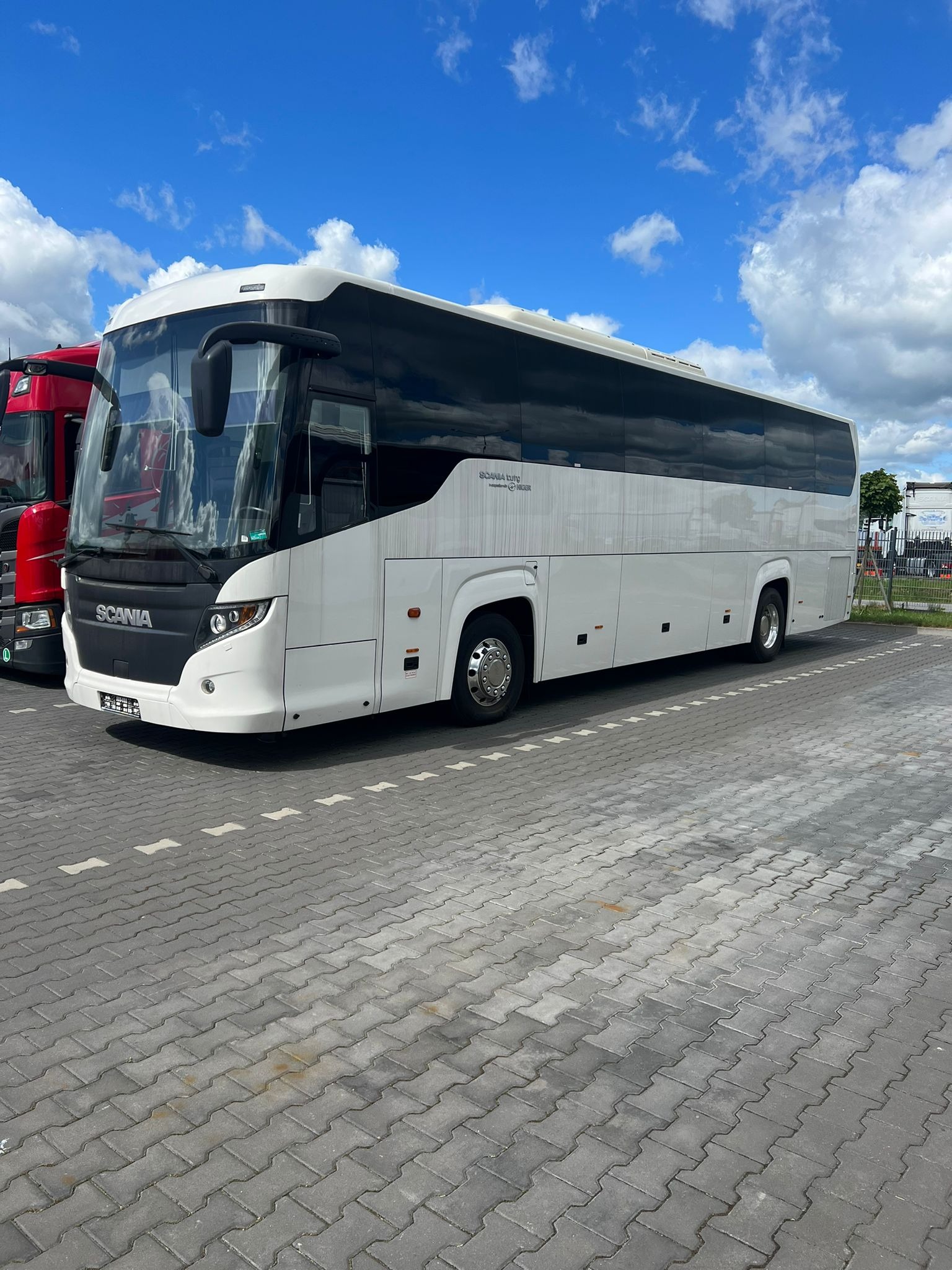 Biały autokar marki Scania zaparkowany na placu z szarej kostki brukowej, obok czerwonej ciężarówki, pod błękitnym niebem z białymi chmurami.