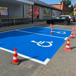 P.H.U. ZNAK Sp.z o.o - Oznakowane na niebiesko miejsce parkingowe dla niepełnosprawnych, wygrodzone pachołkami, na tle parkingu przed sklepem. Oznakowanie poziome.