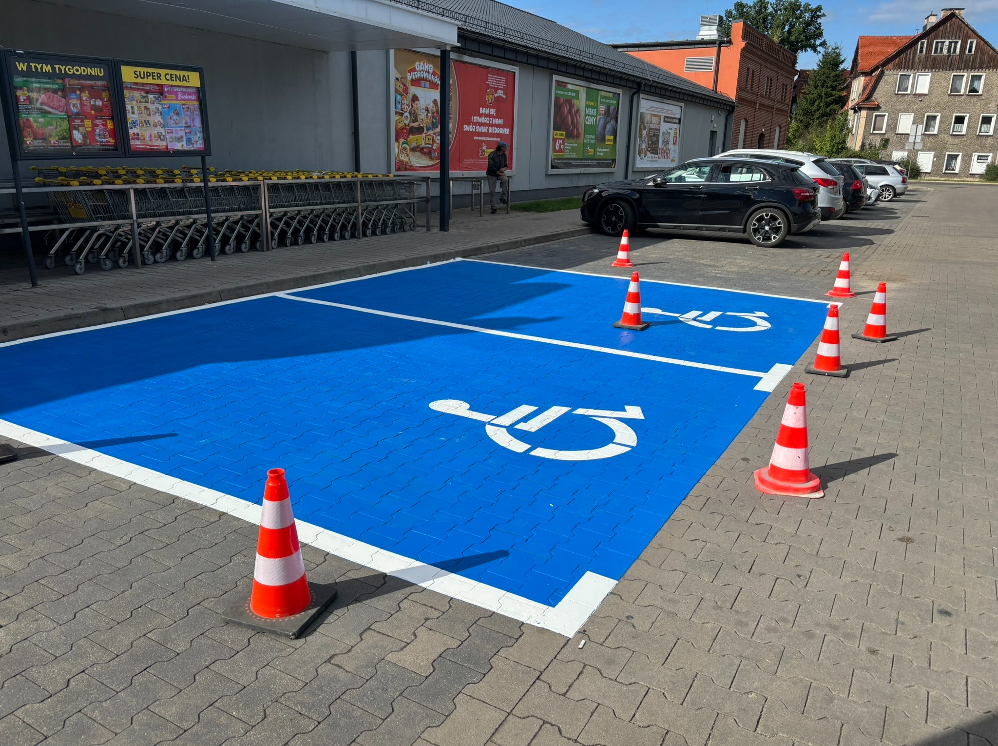 Oznakowane na niebiesko miejsce parkingowe dla niepełnosprawnych, wygrodzone pachołkami, na tle parkingu przed sklepem. Oznakowanie poziome.