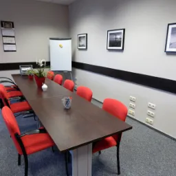 Sala konferencyjna z długim stołem z ciemnego drewna, otoczonym czerwonymi krzesłami, flipchartem, dekoracjami roślinnymi i czarno-białymi zdjęciami na ścianach.