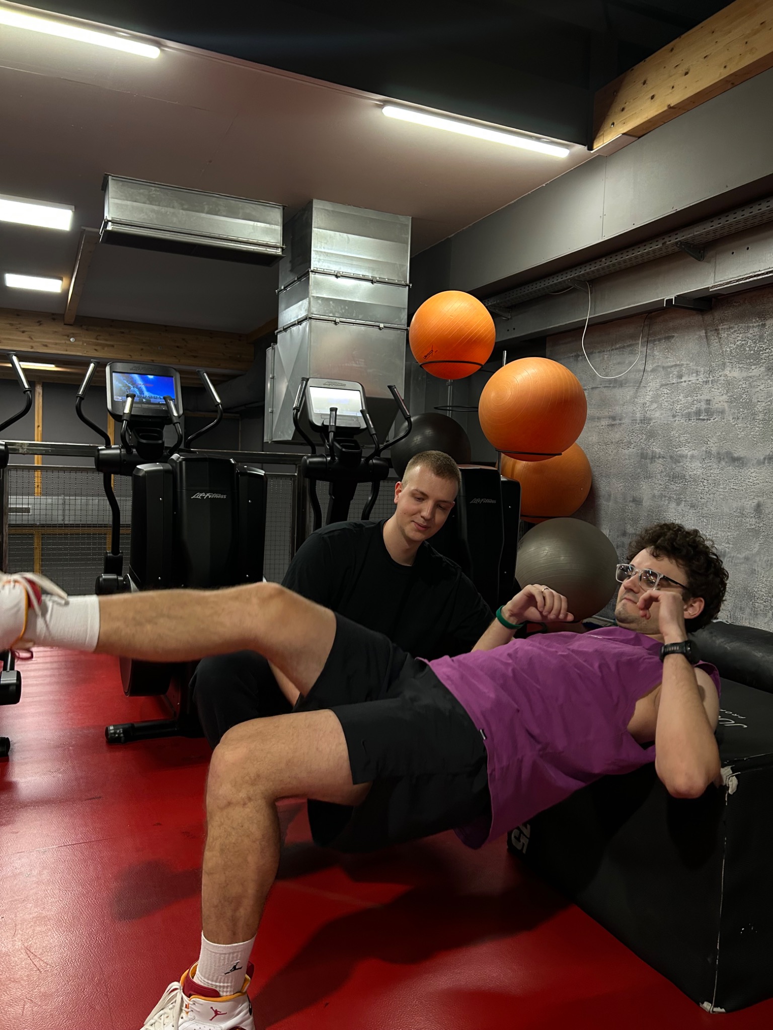 Mężczyzna w okularach leży na czarnej skrzyni w siłowni, unosząc nogę, podczas gdy drugi mężczyzna siedzi obok niego i obserwuje.