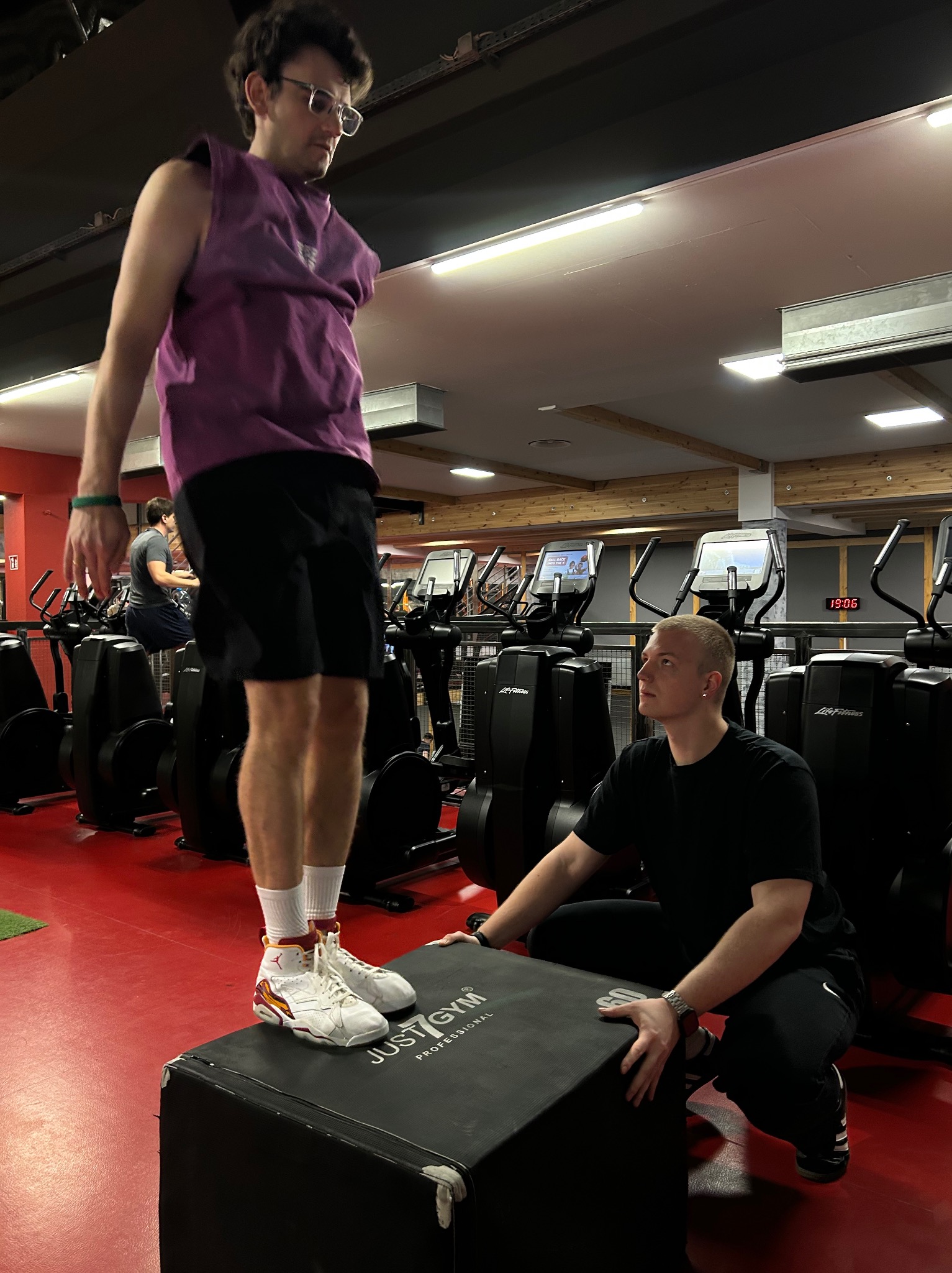 Mężczyzna w fioletowej koszulce i czarnych spodenkach wykonuje ćwiczenie na skrzyni w siłowni, pod nadzorem drugiego mężczyzny w czarnym ubraniu, z widocznymi maszynami do ćwiczeń w tle.