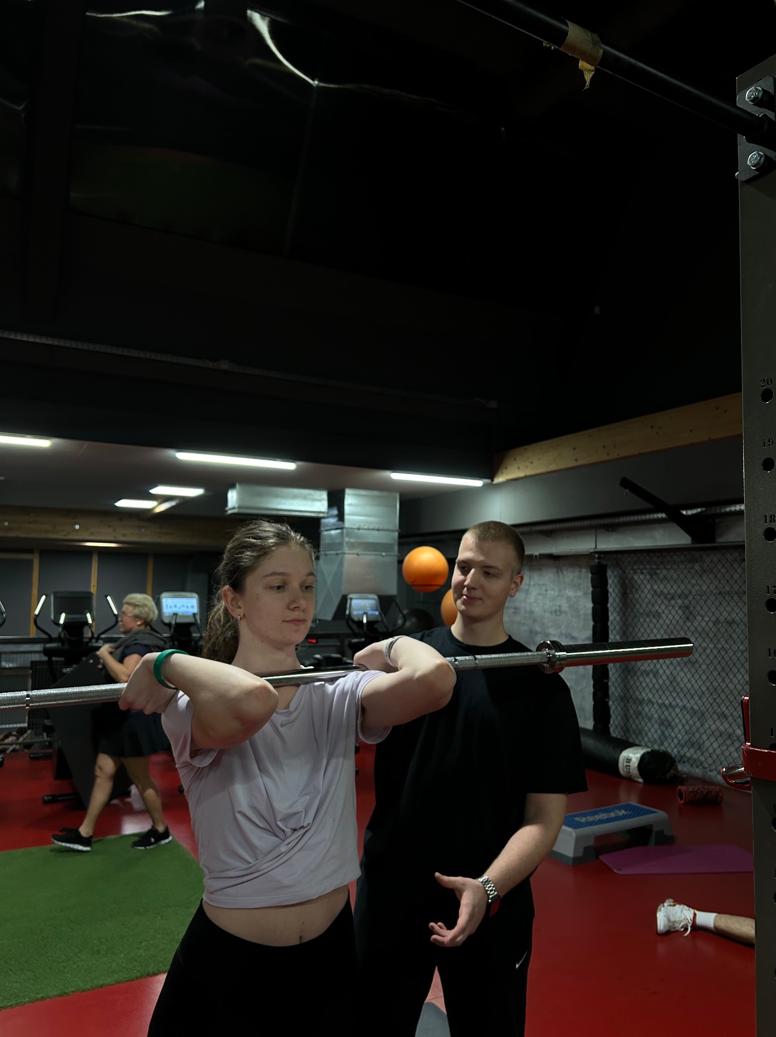 Kobieta ćwiczy podnoszenie sztangi pod nadzorem trenera personalnego w siłowni z czerwono-zieloną podłogą.