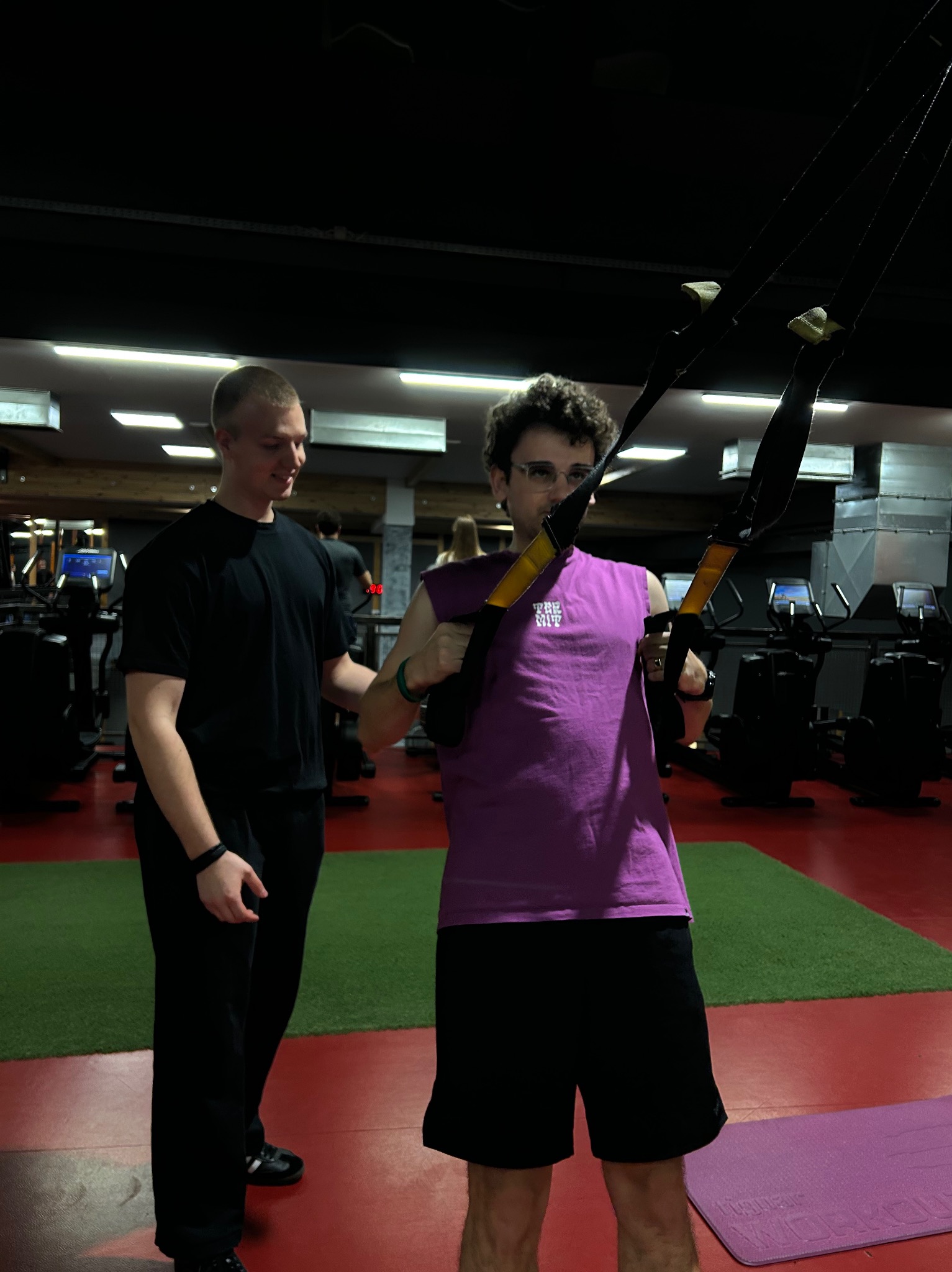 Mężczyzna w okularach i fioletowej koszulce ćwiczy na TRX pod okiem trenera w siłowni z czerwono-zieloną podłogą i sprzętem do ćwiczeń w tle.