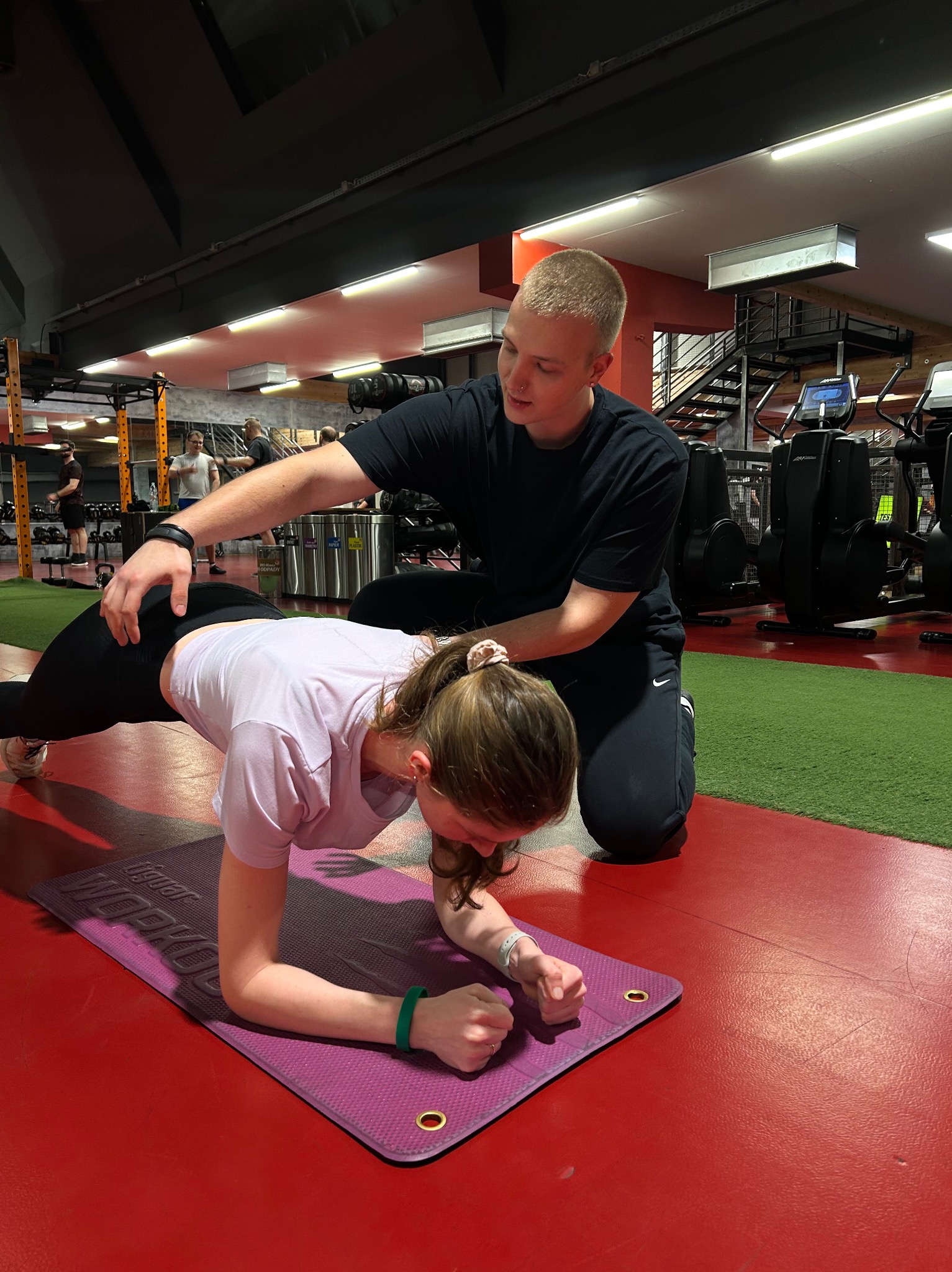 Kobieta wykonuje ćwiczenie plank na fioletowej macie w siłowni, nadzorowana przez mężczyznę w czarnym stroju sportowym, który koryguje jej postawę.