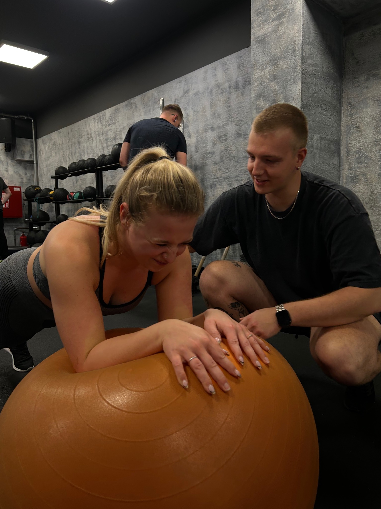 Kobieta w sportowym stroju wykonuje ćwiczenie na pomarańczowej piłce gimnastycznej pod nadzorem mężczyzny w siłowni.