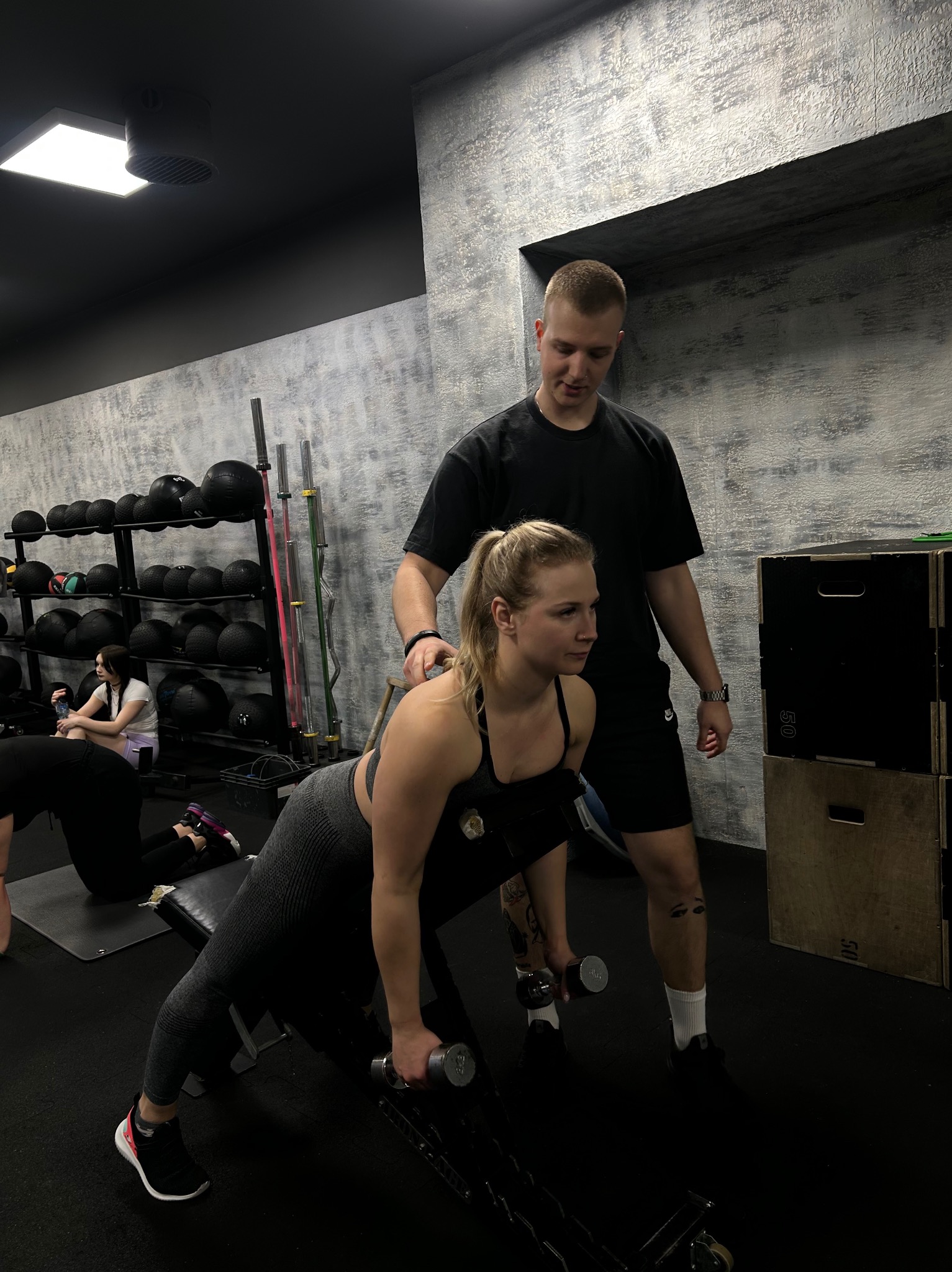 Kobieta w sportowym stroju wykonuje ćwiczenie z hantlami na ławce, a mężczyzna w czarnym stroju asekuruje ją, stojąc za nią w siłowni.