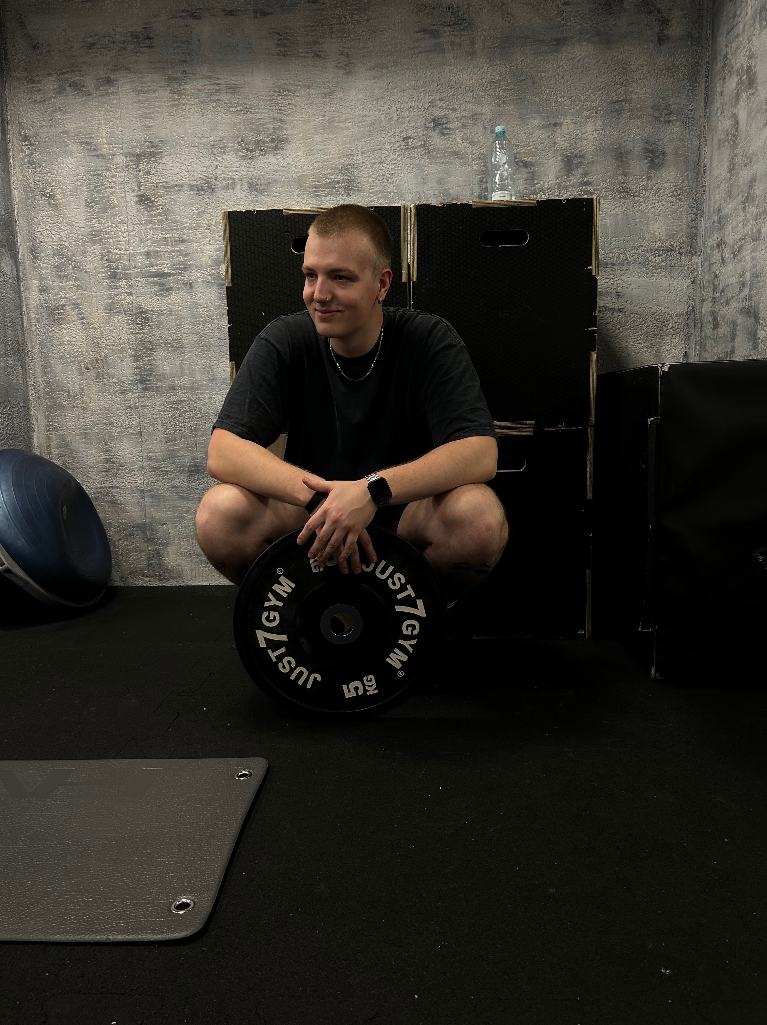 Młody mężczyzna w przysiadzie trzymający obciążenie Just7Gym w siłowni, w tle drabinka gimnastyczna i butelka wody.