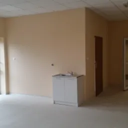 Wnętrze nowo wykończonego pomieszczenia z beżowymi ścianami, jasną podłogą, umywalką z szafką i dwoma parami drzwi, w tym jedne z wyjściem na balkon.