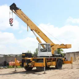 Żółty dźwig samochodowy z wysięgnikiem ustawiony na placu budowy, widoczne podpory stabilizujące i betonowy mur w tle.