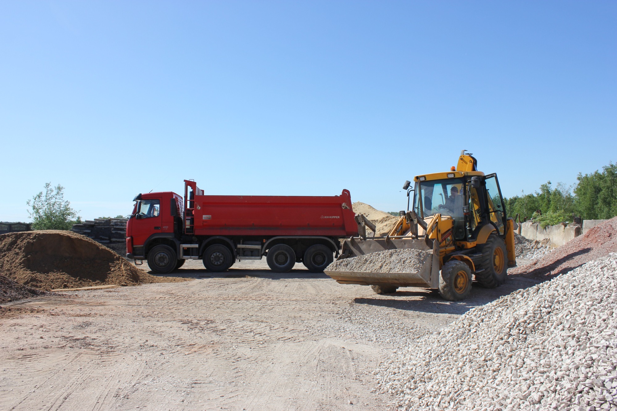 Czerwona wywrotka i żółta koparko-ładowarka na placu budowy, z hałdami piasku i kamieni w tle, błękitne niebo.