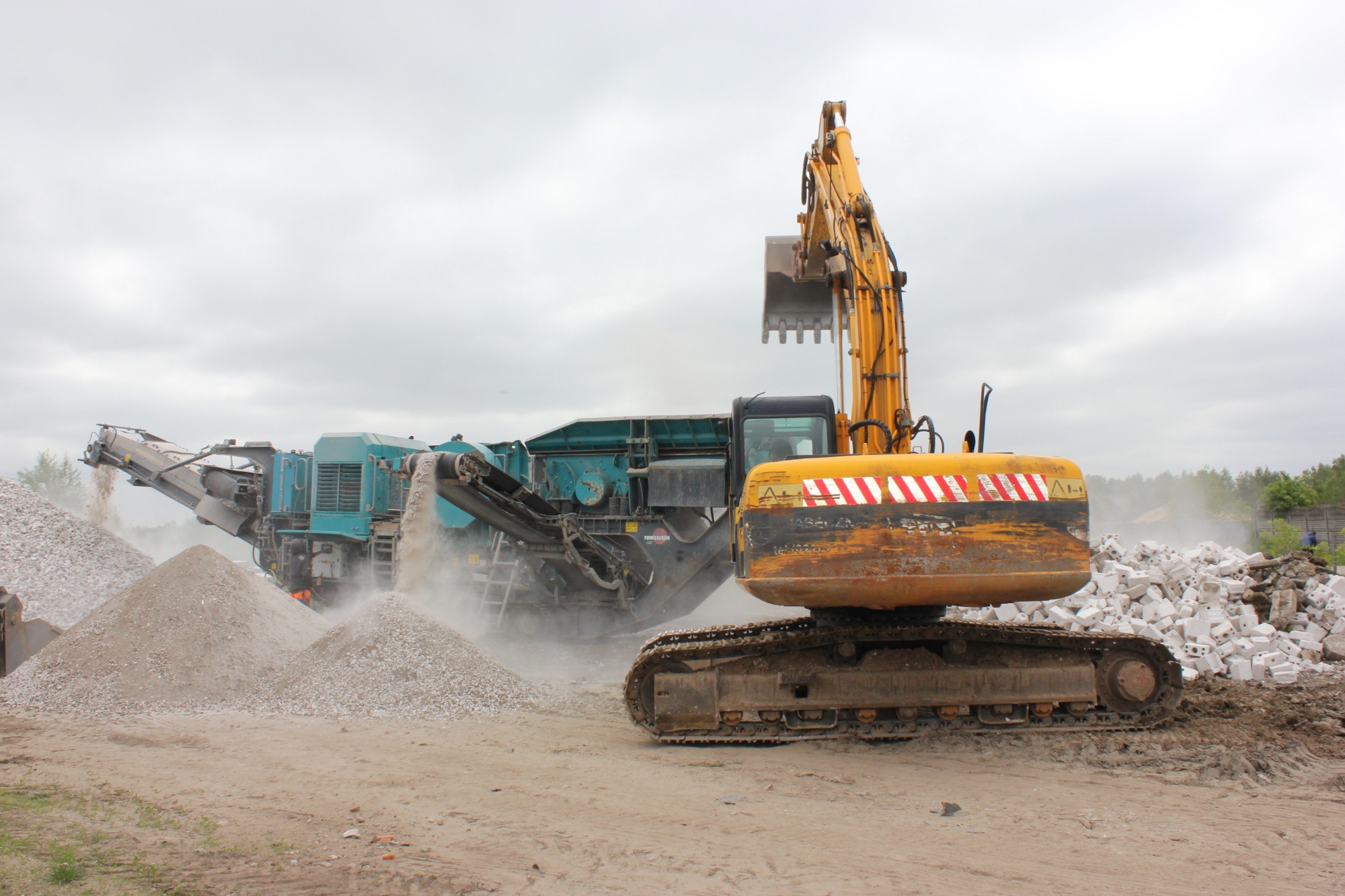 Żółta koparka gąsienicowa z podniesioną łyżką na placu budowy, w tle niebieska kruszarka przesiewająca gruz, tworząca hałdy kruszywa i stertę białych bloków.