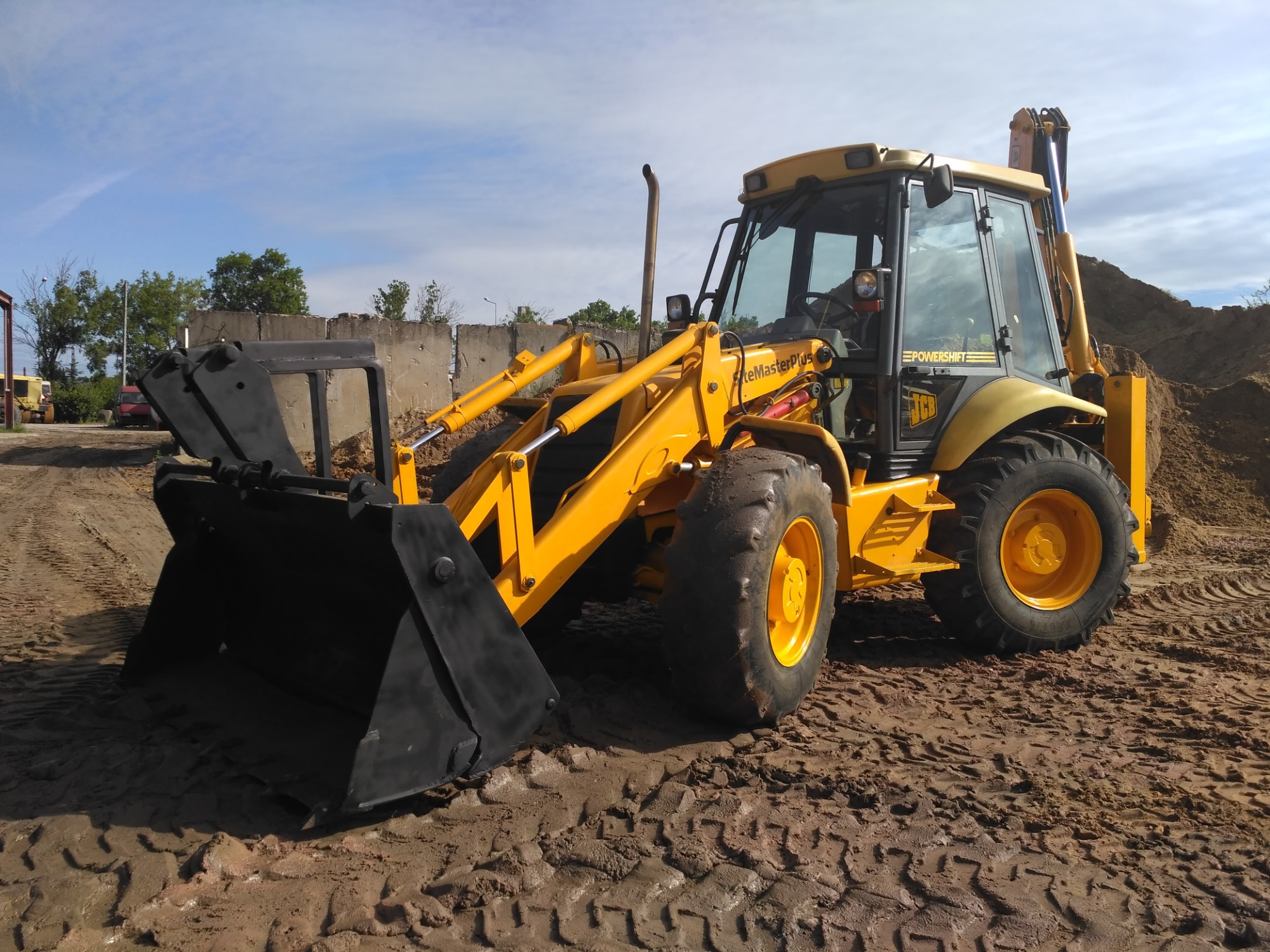 Żółto-czarna koparko-ładowarka JCB SiteMasterPlus na placu budowy z wyraźnymi śladami opon w błotnistym terenie, widoczny stos piasku w tle i fragment betonowego muru.