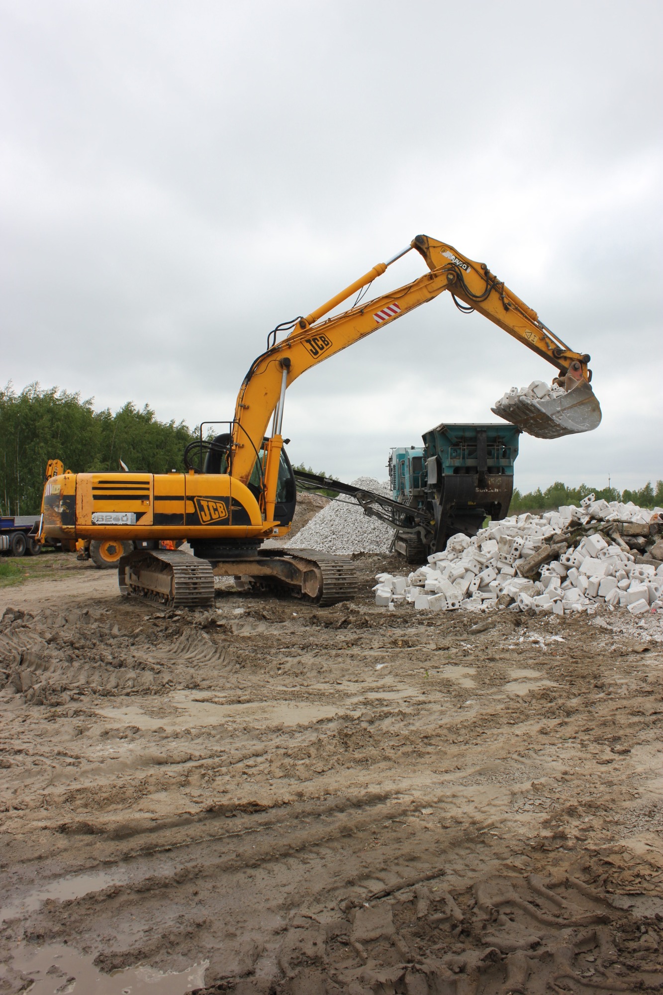 Żółta koparka JCB podnosi gruz i wrzuca go do kruszarki na błotnistym placu budowy, w tle widoczny las pod pochmurnym niebem.