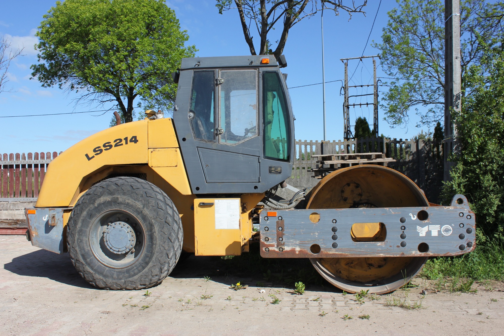 Żółto-szary walec drogowy LSS214 na piaszczystym podłożu, widoczny z boku, z drewnianym płotem i zielenią w tle.