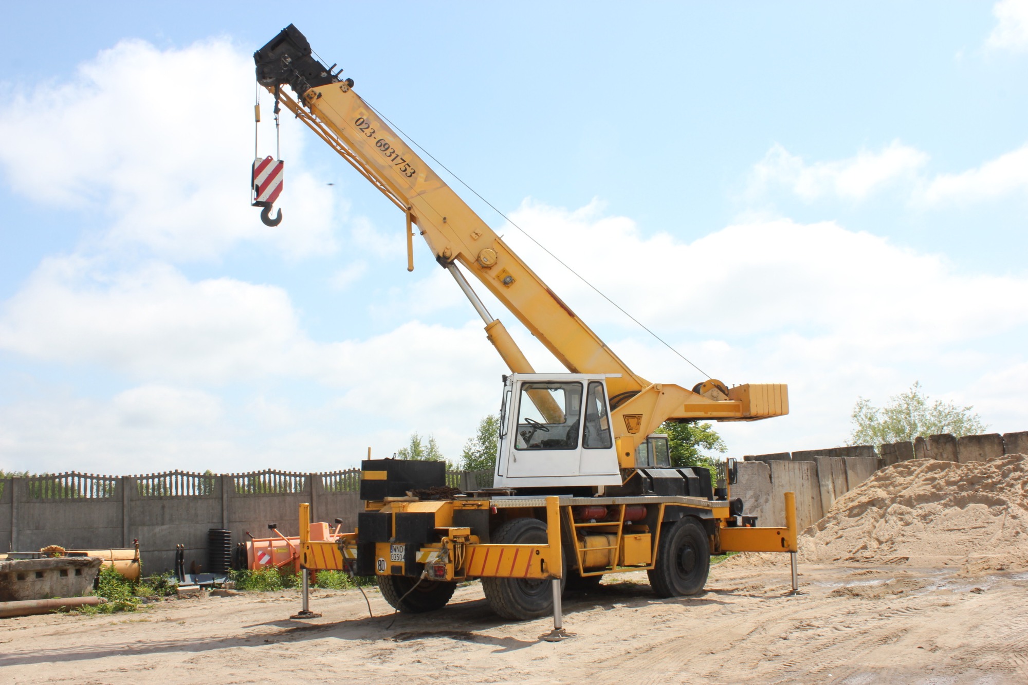 Żółty dźwig samochodowy z wysięgnikiem ustawiony na placu budowy, widoczne podpory stabilizujące i betonowy mur w tle.