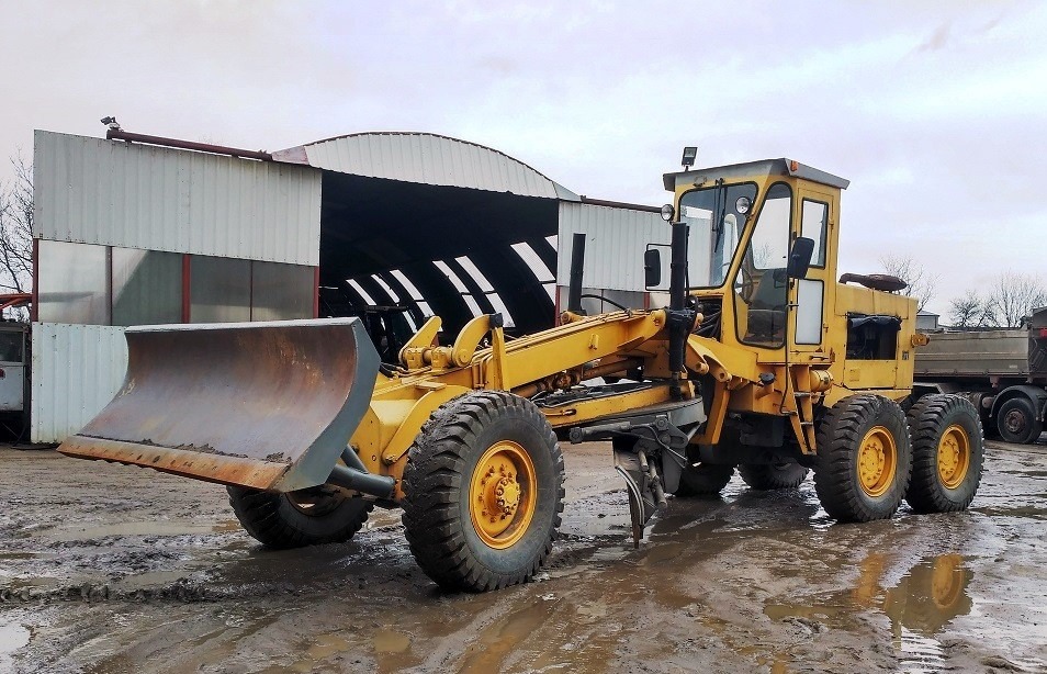 Żółty równiarka drogowa stoi na błotnistym terenie przed budynkiem magazynowym z otwartymi wrotami, widoczne ślady opon i kałuże.