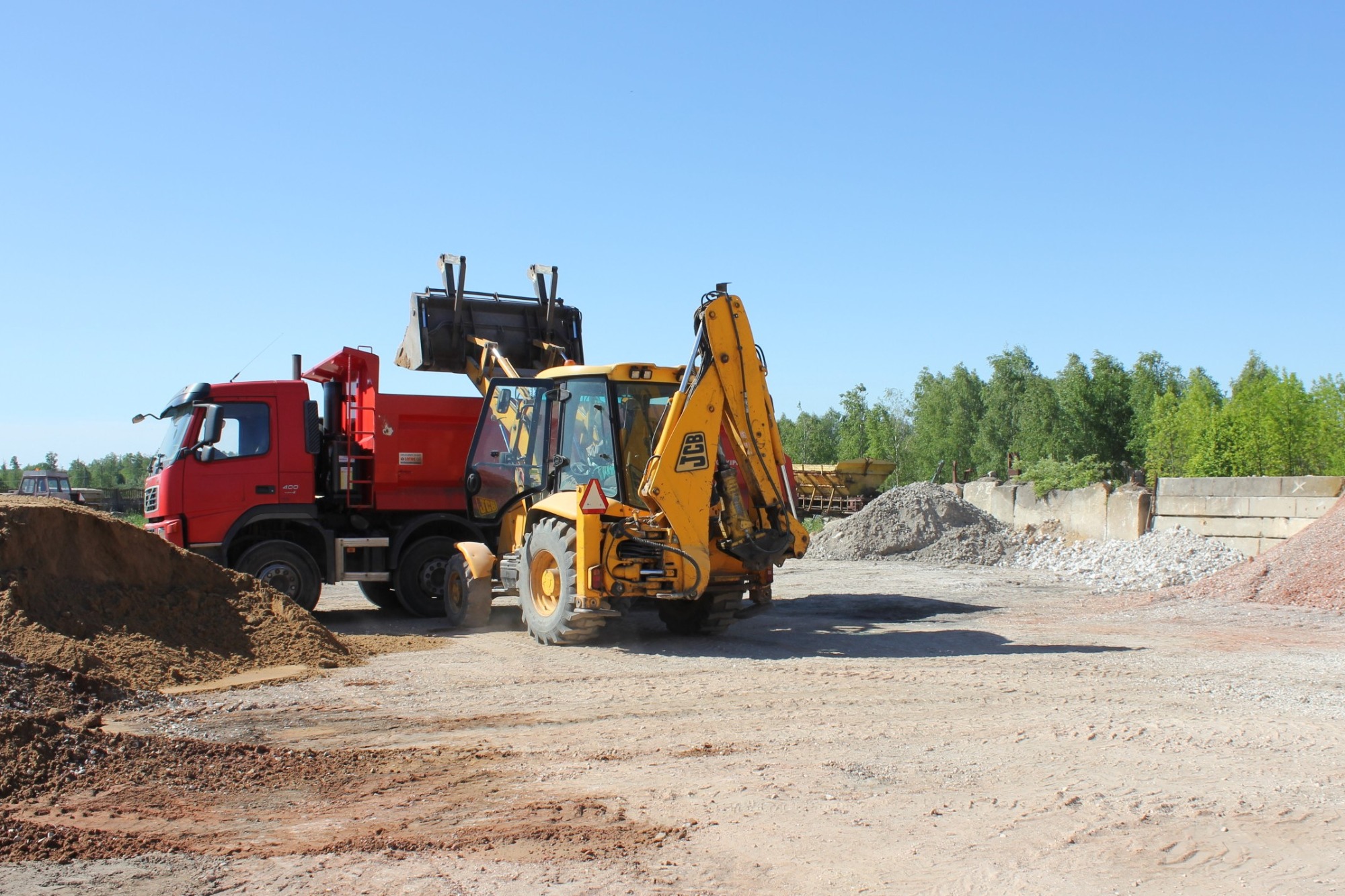 Żółta koparko-ładowarka JCB z podniesioną łyżką, obok czerwona wywrotka z nasypem piasku, na placu budowy z hałdami gruzu i zielenią w tle.