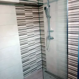 Kabina prysznicowa ze szklaną ścianką, chromowaną armaturą i odpływem liniowym, wykończona płytkami w odcieniach szarości i bieli z poziomymi paskami.