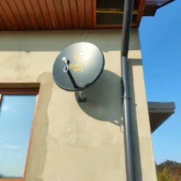 Czarna antena satelitarna z logo Polsat Box zamontowana na jasnej ścianie budynku obok okna i rynny, widoczne drewniane podbitki dachu.