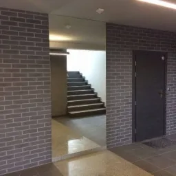 Ściana wyłożona szarą cegłą klinkierową w korytarzu z widocznymi schodami i drzwiami z numerem 10, oświetlona listwą LED na suficie.