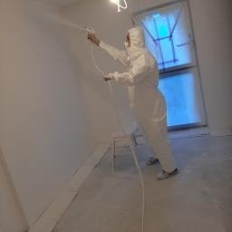 Mal-bud - Pracownik w kombinezonie ochronnym aplikuje białą farbę natryskową na ścianę w pomieszczeniu. Widoczne ślady farby na podłodze i parapecie okiennym.