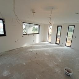 Mal-bud - Wnętrze w stanie deweloperskim przed malowaniem, widoczne ściany po nałożeniu gładzi, surowa podłoga, wystające przewody elektryczne i czarne ramy okienne.