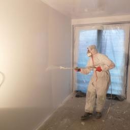 Pracownik w kombinezonie ochronnym i masce natryskuje gładź na ścianę w pomieszczeniu zabezpieczonym folią malarską, widoczny wąż i dysza urządzenia.