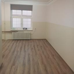 Pusty pokój po remoncie: panele podłogowe imitujące drewno, biało-beżowe ściany z widocznym podziałem kolorystycznym, grzejnik żeliwny pod oknem z szarymi roletami. Widoczne gniazdko elektryczne.