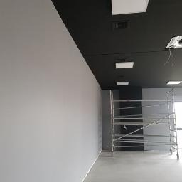 Świeżo pomalowane na szaro ściany w biurze, czarny sufit z zamontowanym oświetleniem, widoczne rusztowanie na kółkach, betonowa podłoga.