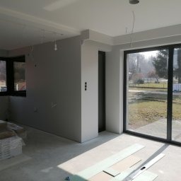 Szare ściany w surowym, jasnym wnętrzu z dużym oknem przesuwnym i pakietem płyt gipsowo-kartonowych na podłodze w rogu pomieszczenia.