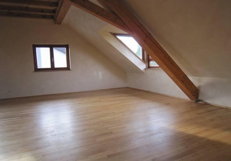 Poddasze po remoncie: jasna drewniana podłoga, gładkie ściany w odcieniach beżu, drewniane belki stropowe i okna dachowe w brązowych ramach.