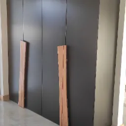 Duża, matowa, grafitowa szafa wnękowa z nietypowymi, wysokimi, drewnianymi uchwytami, widoczna na tle jasnej ściany i podłogi z płytek.