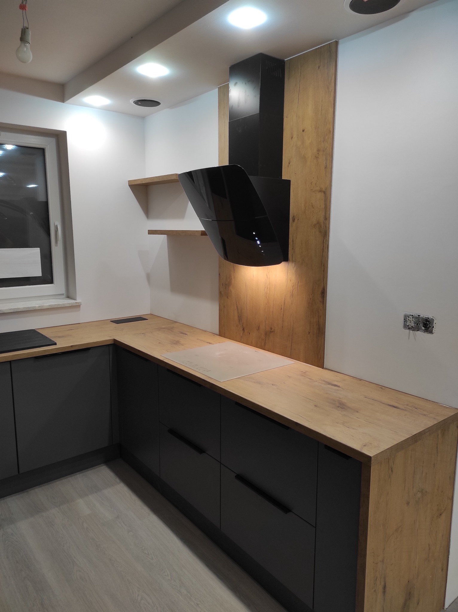 Nowoczesna kuchnia z szarymi szafkami i blatem imitującym drewno, czarny okap o futurystycznym kształcie, oświetlenie punktowe i półki wiszące.