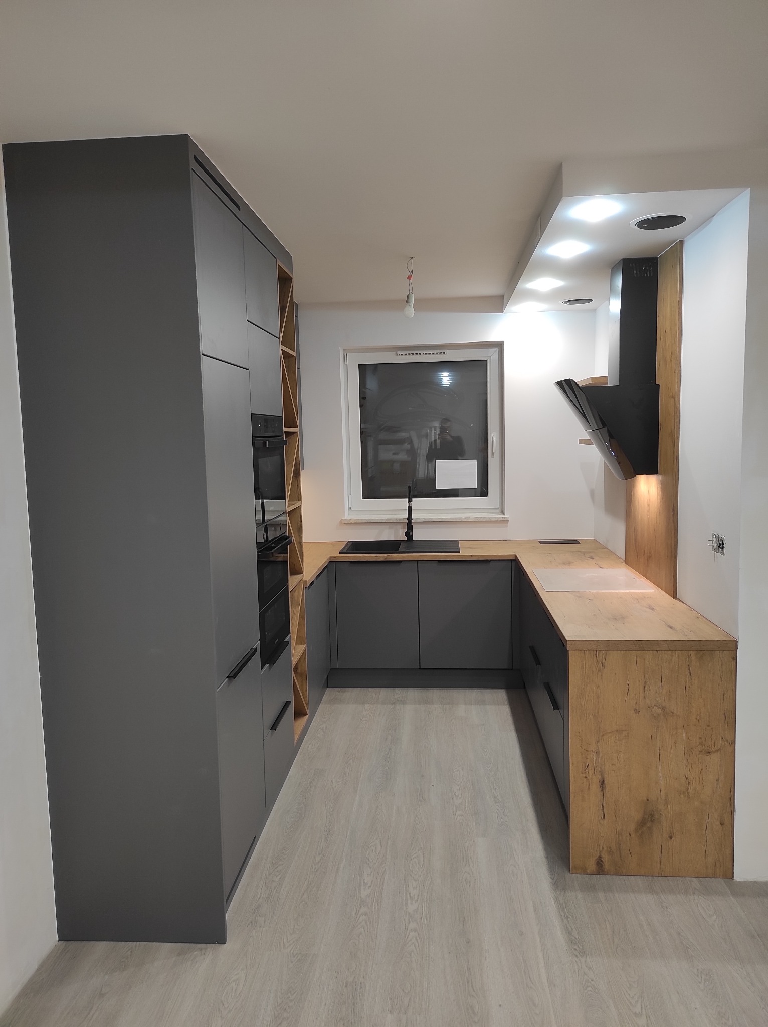 Nowoczesna kuchnia w odcieniach szarości z drewnianym blatem i zabudową piekarnika, oświetleniem punktowym nad blatem i czarnym okapem.