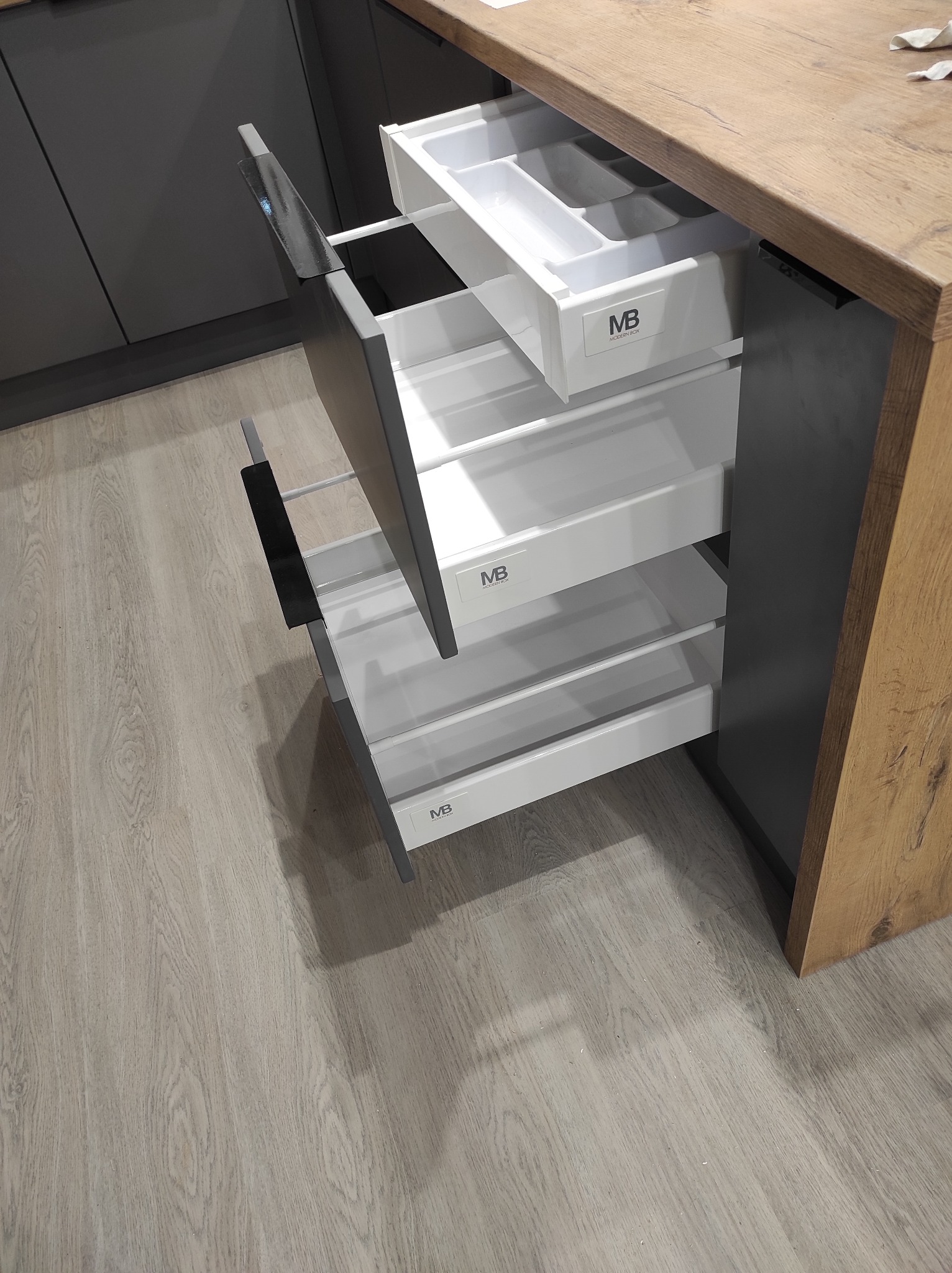 Wysuwane, białe szuflady z organizerem w nowoczesnej, szarej kuchni z blatem imitującym drewno, widoczne logo producenta na frontach szuflad.