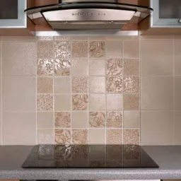 Beżowe kwadratowe płytki z różnymi wzorami kwiatowymi i teksturowanymi, ułożone na ścianie nad płytą indukcyjną w kuchni, z widocznym okapem i fragmentami szafek.