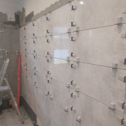 ADBUD Adam Bałdowski - Ściana w trakcie układania płytek z użyciem krzyżyków dystansowych, widoczna drabina i poziomica.