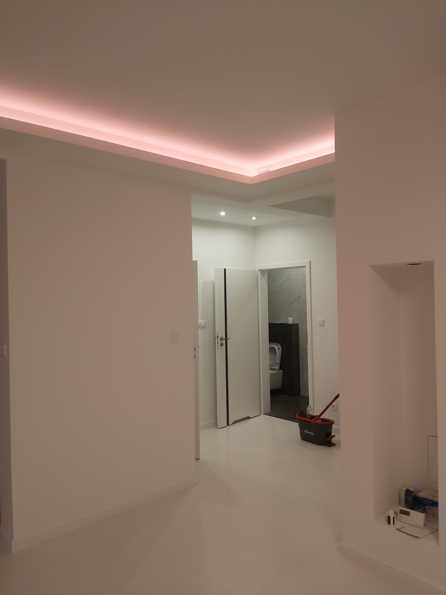 Wnętrze po remoncie: białe ściany, podłoga z połyskiem, podświetlany sufit i widok na wykończoną łazienkę z marmurowymi płytkami