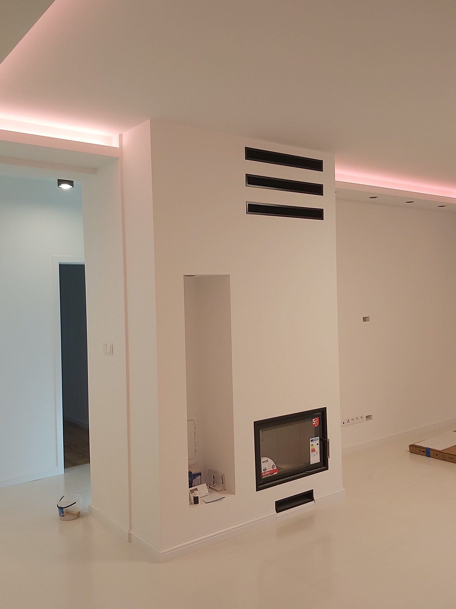 Minimalistyczne wnętrze w Ludwinowie Dębskim po remoncie, z białymi ścianami, podłogą i zabudową kominka z wkładem kominkowym oraz dekoracyjnym oświetleniem LED przy suficie.