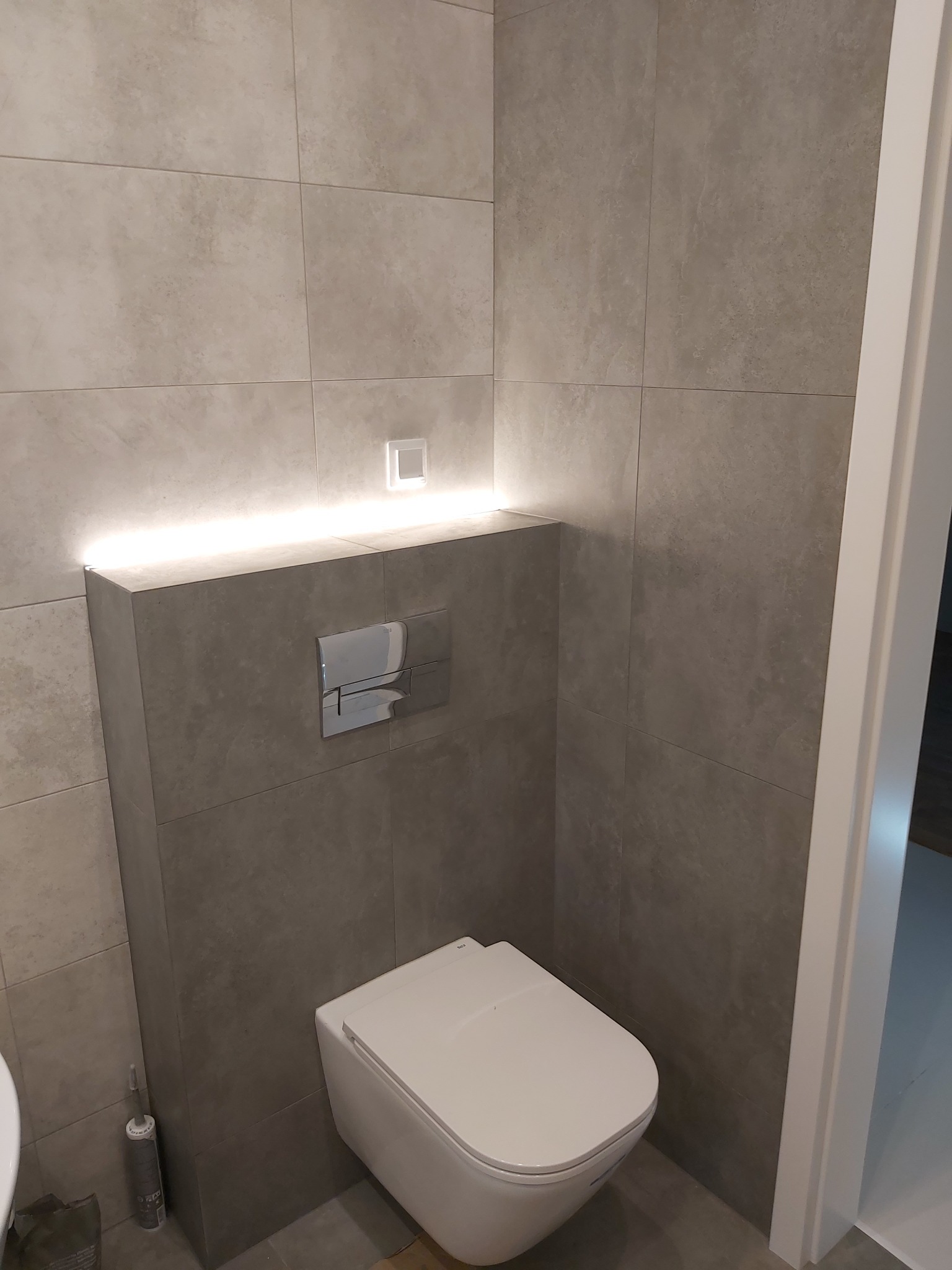 Nowoczesna toaleta podwieszana z szarymi płytkami i podświetleniem LED nad zabudową stelaża.