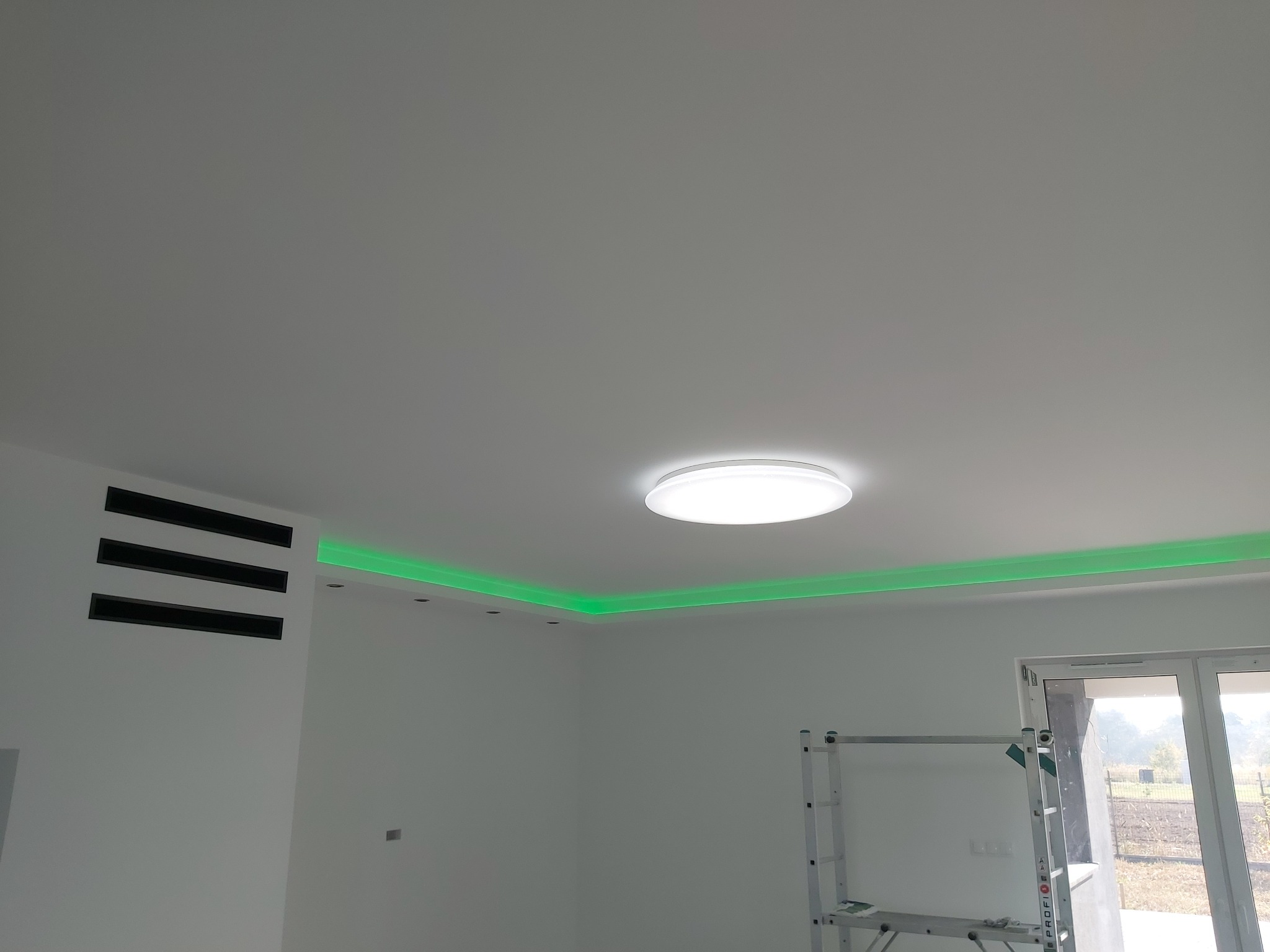 Białe wnętrze z podświetlonym na zielono sufitem LED i okrągłą lampą sufitową, widoczna drabina malarska w rogu.