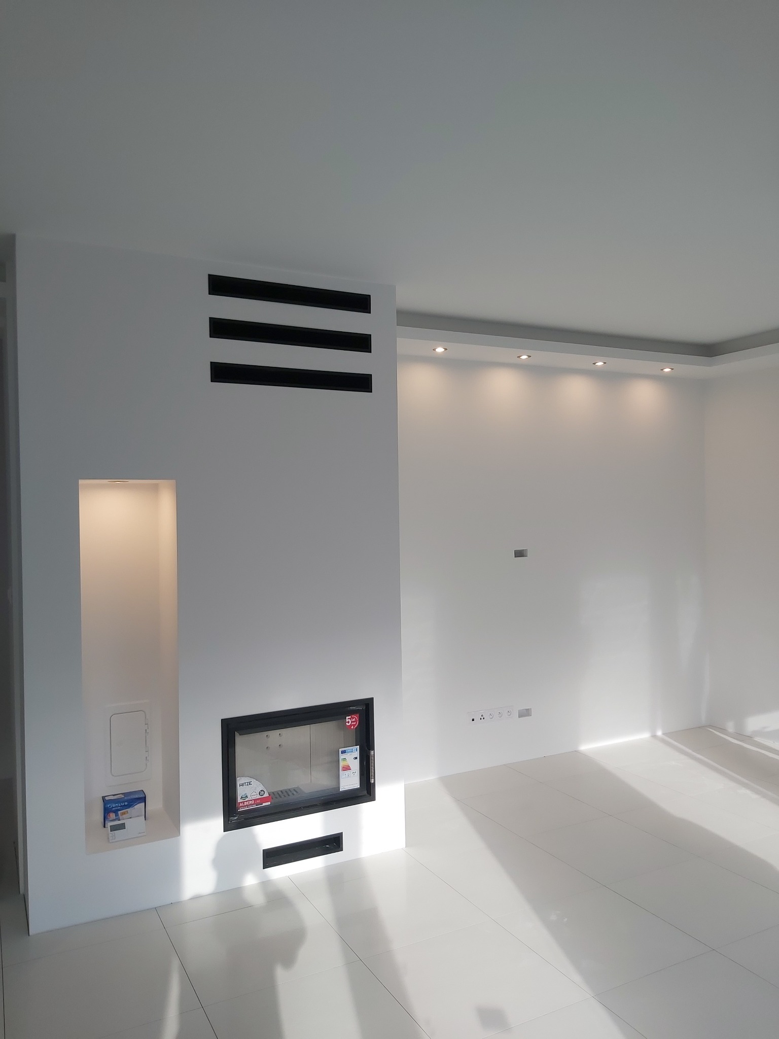 Minimalistyczne wnętrze z białymi płytkami podłogowymi, wbudowanym kominkiem z czarną ramą, trzema poziomymi czarnymi paskami nad kominkiem i wnęką z półką, oświetlone punktowo.