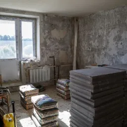 Wnętrze pokoju w trakcie prac przygotowawczych: surowe ściany, okno z widokiem na zieleń, grzejnik, worki z zaprawą i stos płyt.