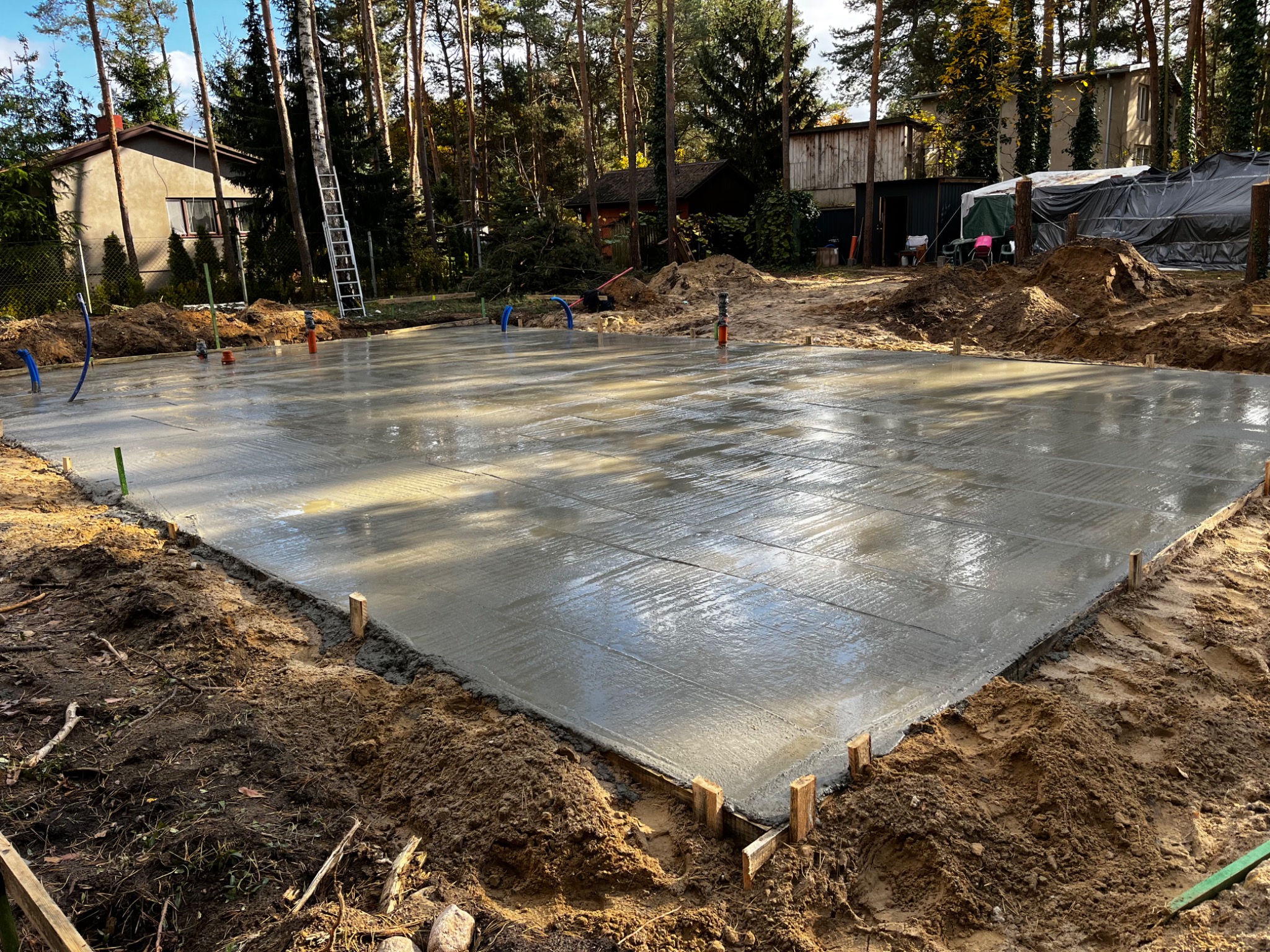 Świeżo wylana, mokra płyta fundamentowa na gruncie, otoczona drewnianym szalunkiem i wystającymi rurami instalacyjnymi, na tle zalesionego terenu.