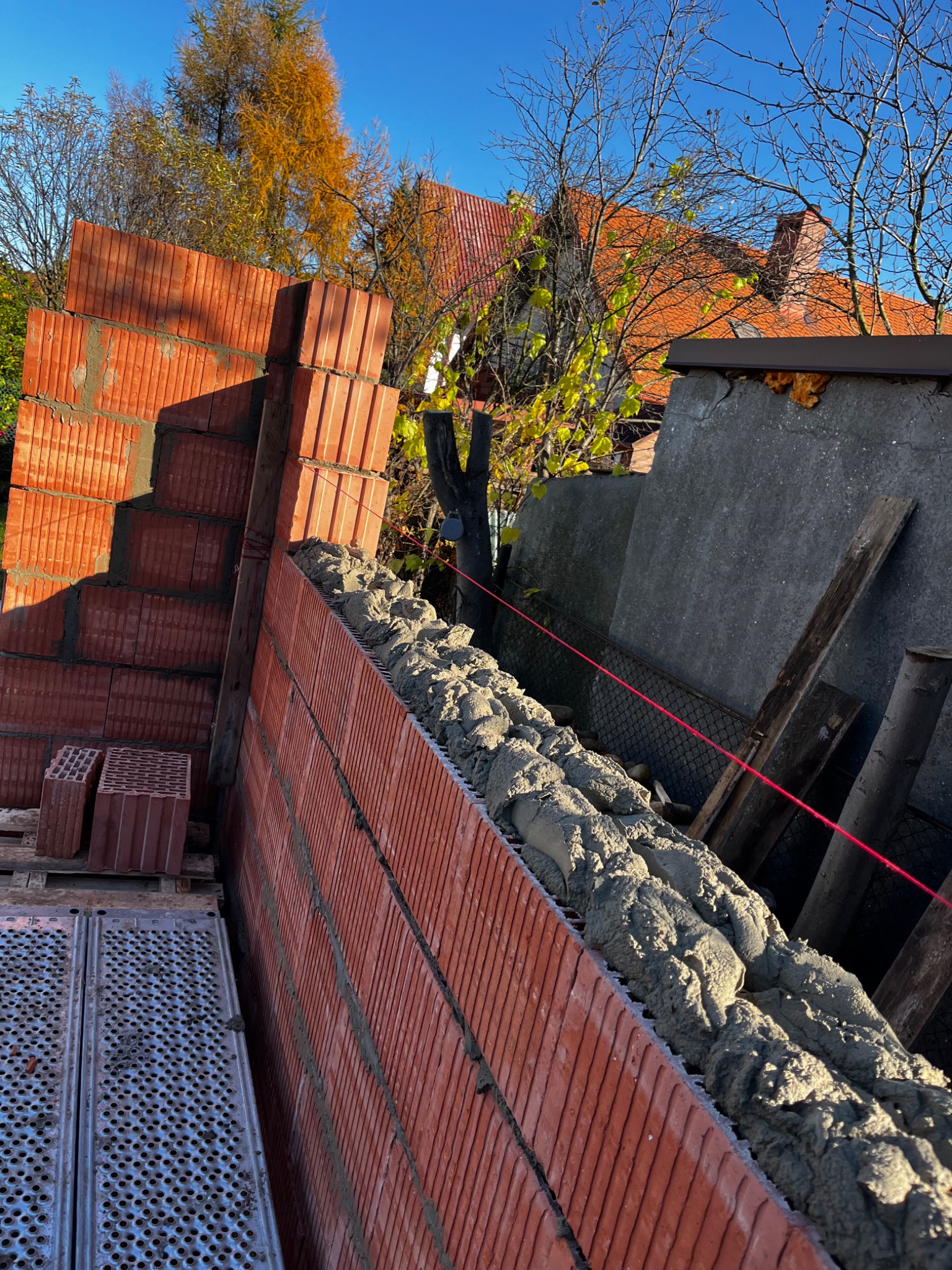 Wznoszenie ściany z czerwonej cegły szczelinowej na zaprawie, widoczne rusztowanie i poziomica laserowa, w tle budynek z czerwoną dachówką i drzewa z jesiennymi liśćmi.