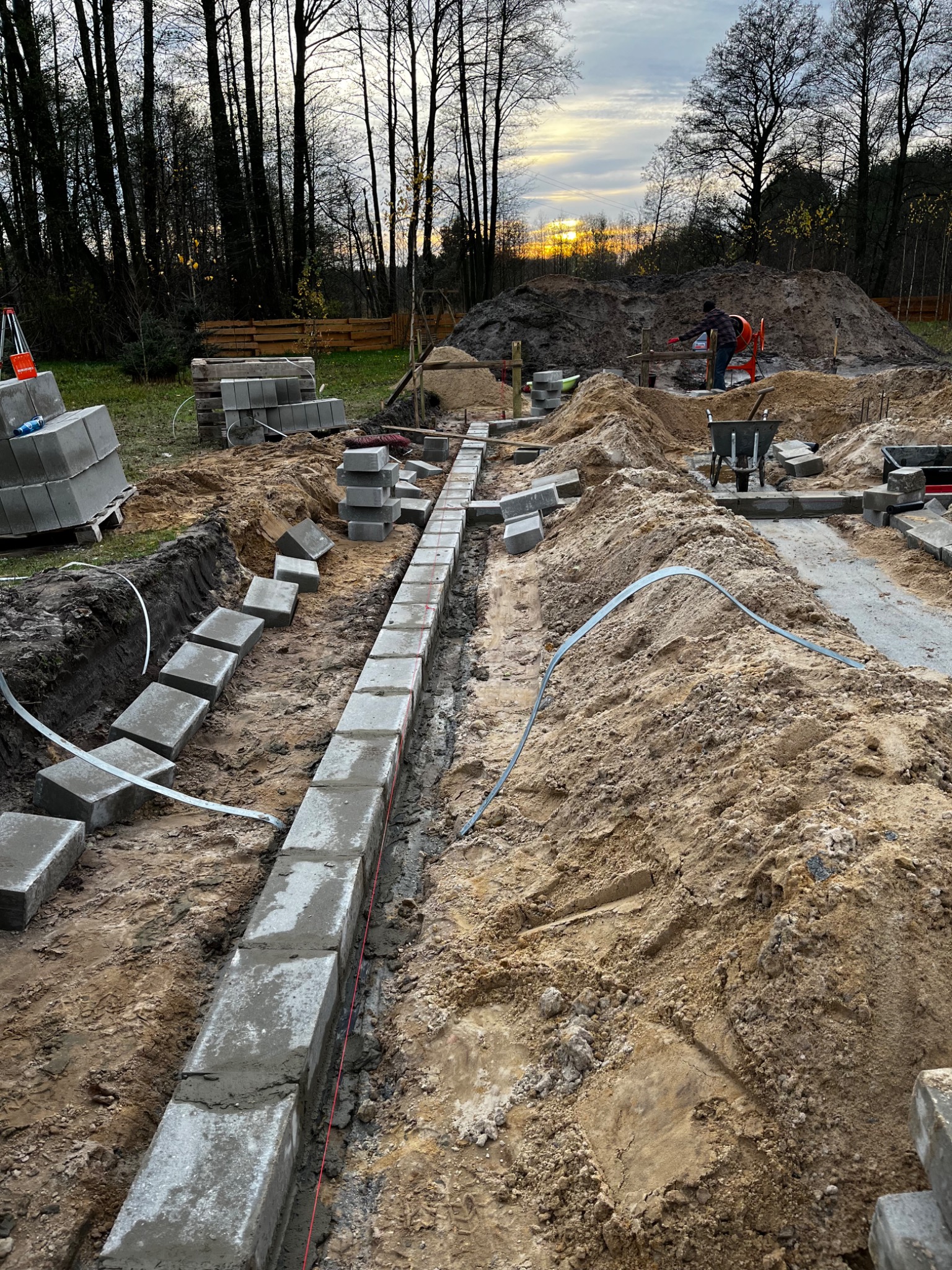 Wykop pod fundament z ułożonymi betonowymi bloczkami, widoczny pracownik przy mieszalniku i zachodzące słońce w tle.